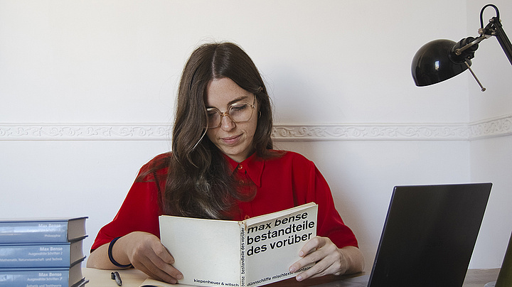 Rosa Coppola, eine junge Frau mit langen braunen Haaren, die eine Brille und ein rotes Hemd trägt, sitzt an einem Schreibtisch. Sie liest ein Buch mit dem Titel „Max Bense – Bestandteile des Vorüber“, neben ihr stehen ein Laptop und ein Stapel Bücher. In der Ecke ist eine schwarze Schreibtischlampe zu sehen.