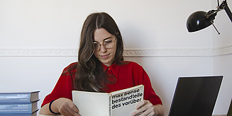 Rosa Coppola, eine junge Frau mit langen braunen Haaren, die eine Brille und ein rotes Hemd trägt, sitzt an einem Schreibtisch. Sie liest ein Buch mit dem Titel „Max Bense – Bestandteile des Vorüber“, neben ihr stehen ein Laptop und ein Stapel Bücher. In der Ecke ist eine schwarze Schreibtischlampe zu sehen.